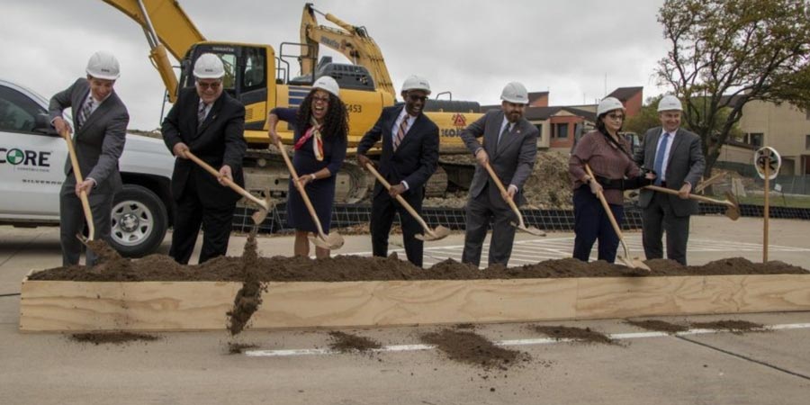 The photo depicts seven college and community leaders in a row, holding shovels and wearing construction hard hats. They are all smiling and happy, digging into the dirt and celebrating the joyous occasion to start the construction on a new building for the Eastfield Campus that will serve students and the community.  Behind the seven people, is a large construction bull dozer and a pick-up truck.